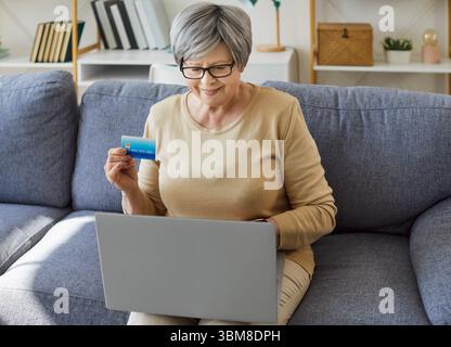 Donna anziana felice seduta sul divano con laptop, con carta di credito mentre fa acquisti online a casa. Foto Stock
