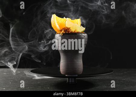 Ciotola di narghilè con agrumi e fumo sul tavolo nero, primo piano Foto Stock