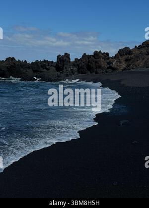 Spiaggia di sabbia nera con dolci onde, scogliere rocciose sullo sfondo e cielo azzurro. Tranquilla scena costiera caratterizzata da una sabbia nera vulcanica unica. Foto Stock