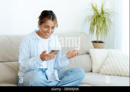 La felice donna brasiliana si siede sul divano, ridendo e chiacchierando durante le videochiamate sullo smartphone. Rilassata ama passare il tempo scorrendo i social media e leggendo buone notizie a casa Foto Stock