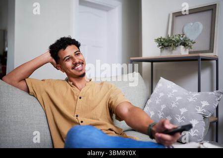 Uomo sorridente che si rilassa sul divano tenendo in mano il telecomando in un accogliente soggiorno Foto Stock