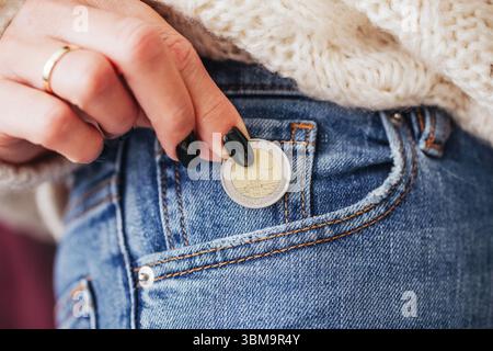 Donna che mette la moneta nella tasca dei jeans. Mano che tiene in mano denaro in euro. Concetto di risparmio e finanziamento. Economia personale e budget. Primo piano delle dita. Foto Stock