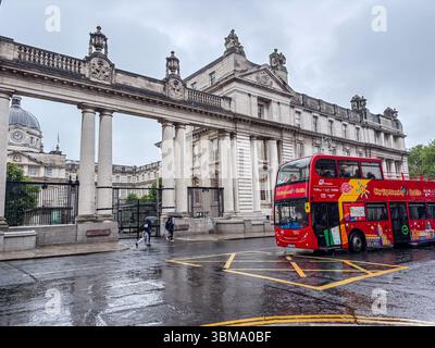 Edifici governativi a Dublino in una giornata di pioggia con un autobus rosso a due piani e persone con ombrelloni. Foto Stock