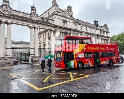 L'autobus rosso a due piani City Sightseeing passa davanti a un edificio storico durante una giornata di pioggia a Dublino, Irlanda. I pedoni con ombrelli sono visibili. Foto Stock
