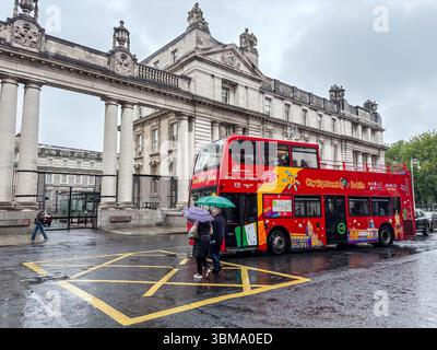 Autobus a due piani City Sightseeing a Dublino durante una giornata di pioggia. I pedoni con ombrelloni camminano sulla strada bagnata vicino a un grande edificio storico. Foto Stock