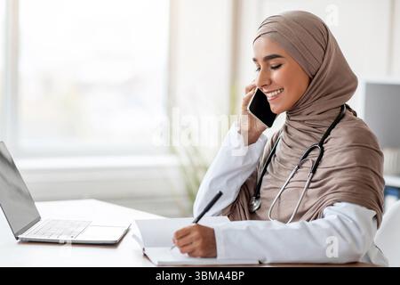 Signora in hijab medico parlare al telefono e prendere appunti Foto Stock