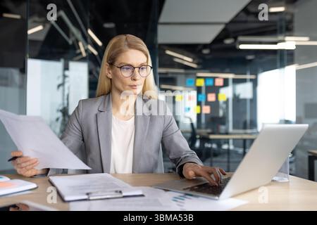 Una donna d'affari lavora su un notebook in un ufficio moderno, rivedendo i documenti mentre lavora. Foto Stock