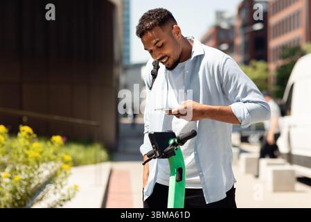 Servizi di condivisione scooter elettrici push. Giovane ragazzo nero che noleggi e-scooters in città, pagando online con smartphone Foto Stock