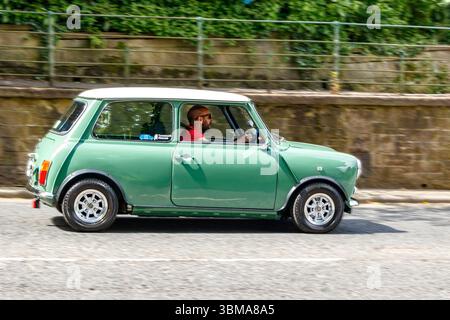 Una classica Mini Cooper verde con tetto bianco è guidata da un uomo in una giornata di sole, che viaggia lungo un muro di pietra vicino al verde. Foto Stock