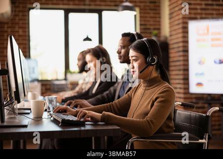 Professionista femminile dedicato in sedia a rotelle, che indossa cuffie cablate, si concentra sulle attività svolte presso la scrivania dell'ufficio. Donna asiatica imprenditrice si impegna con i clienti online, aggiornando i record sul suo computer. Foto Stock