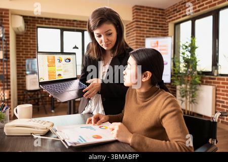 La dipendente caucasica possiede un laptop e riceve consigli strategici dal suo manager asiatico in un ufficio murato. Due giovani donne d'affari collaborano utilizzando strumenti e documenti digitali. Foto Stock