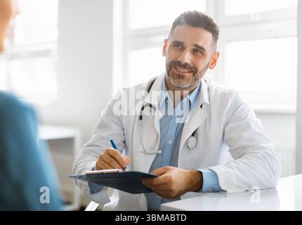 Allegro medico di mezza età in mantello bianco con stetoscopio di consulenza paziente in clinica ufficio interno Foto Stock