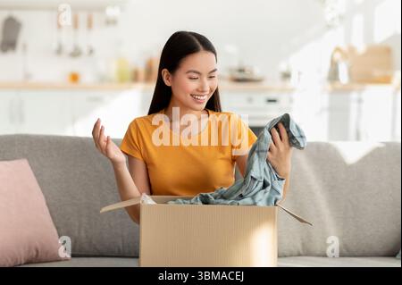 Acquirente soddisfatto dell'acquisto online. Felice donna coreana sboxing pacco, guardando nuova camicia e sorridente Foto Stock