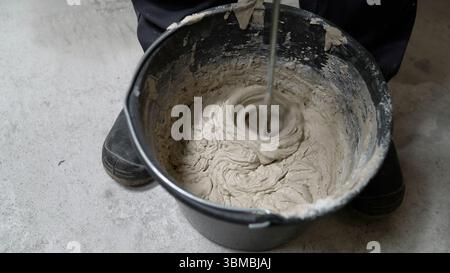 L'operaio edile mescola resina epossidica grigia all'interno di un secchio industriale con una pala di miscelazione specializzata durante il progetto di miglioramento domestico Foto Stock