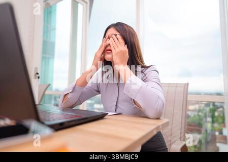 Donna d'affari indonesiana del sud-est asiatico che vive stress e esaurimento mentre lavora su un notebook in un luogo di lavoro ibrido. Scartoffie e un cellulare su Foto Stock