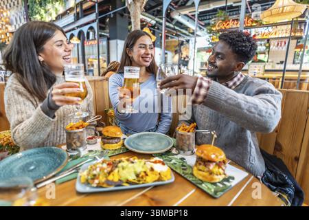 Amici multietnici felici che brindano e gusteranno un pasto delizioso insieme in un ristorante vivace, pieno di risate e gioia Foto Stock
