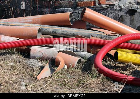 Pile miste di tubi di plastica e metallo arancioni, neri, rossi e gialli, alcune ondulate, giacciono su erba secca accanto a una parete di mattoni, indicando la costruzione Foto Stock