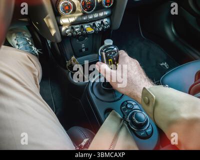Parigi, Francia - 27 marzo 2025: Leva del cambio manuale nell'abitacolo elettrico della Mini Cooper con pantaloni beige pulsanti di comando multipli visibili, indicatori luminosi della climatizzazione e occhiali da sole nel portabicchieri Foto Stock
