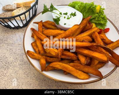 Un piatto di patatine fritte croccanti servite con salsa cremosa e guarnite con lattuga ed erbe aromatiche, accompagnate da un cestino di fette di pane assortite su un tavolo di pietra. Foto Stock