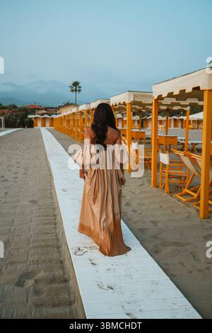 La donna in un abito fluente cammina lungo un sentiero di spiaggia sabbiosa fiancheggiato da tende gialle. L'ambiente sereno riflette un momento di pace in mezzo allo scenario costiero Foto Stock