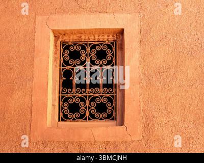 Marocco, Merzouga, 04 luglio 2025. Tradizionale finestra marocchina con griglia ornata in ferro battuto su una parete in adobe, illuminata dal caldo sole pomeridiano nel Sah Foto Stock