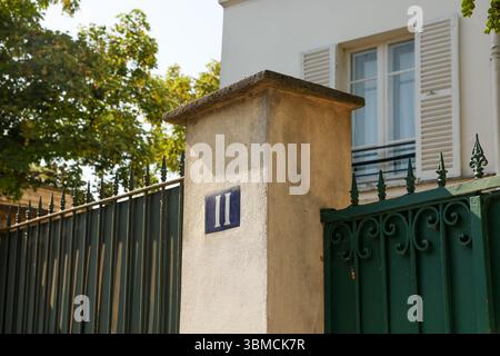 Vista ravvicinata dell'ingresso di una casa caratterizzata da un cancello in metallo verde scuro con decorazioni a scorrimento, una colonna in pietra beige con una piastrella blu numero 'II' e una S. Foto Stock