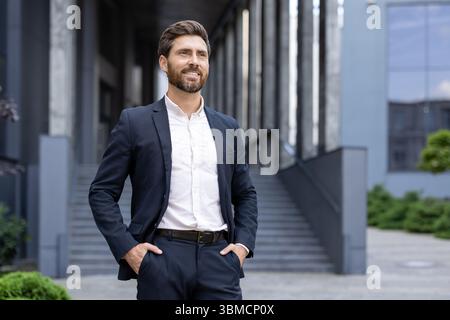Un uomo d'affari sorride mentre si trova all'aperto, vicino a un moderno edificio di uffici, con le mani in tasca. Foto Stock