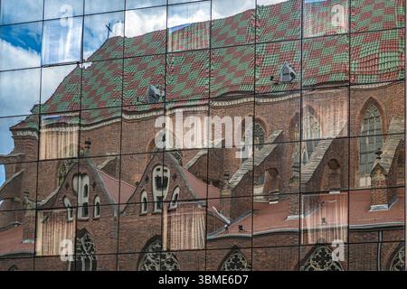 Riflesso di una chiesa storica con un tetto dai motivi vivaci su una moderna facciata in vetro, che mostra contrasti architettonici Foto Stock