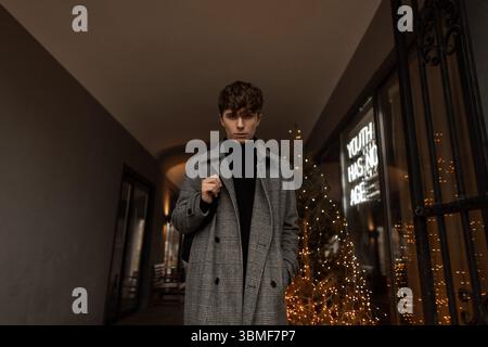 Bellissimo giovane uomo in abiti alla moda autunnali con Uno zaino alla moda in città vicino A Un negozio decorato con luci di Capodanno. Guy FAS Foto Stock