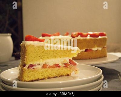 Primo piano della torta di fragole a fette con panna montata in primo piano e la torta completa visibile sullo sfondo. Foto Stock