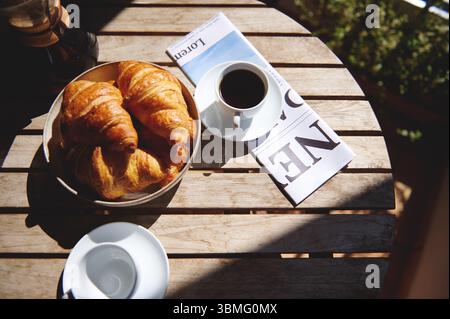 Colazione illuminata dal sole con croissant friabili, caffè nero e quotidiano su un tavolo di legno, che evocano relax e delizie culinarie in un'atmosfera di relax Foto Stock