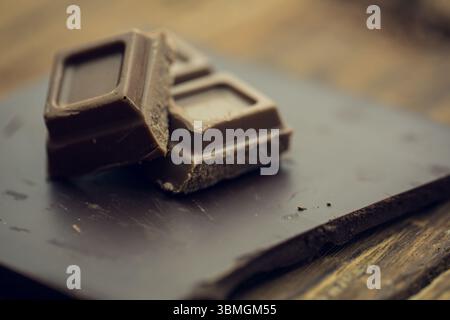 Due quadrati di cioccolato fondente poggiano su una tavola di ardesia su un piano in legno rustico dal design piatto Foto Stock
