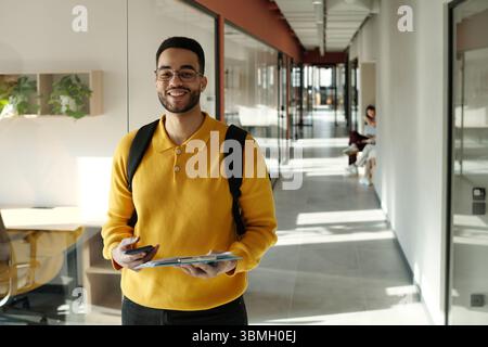 Uomo birazziale con barba che indossa un maglione giallo e occhiali in piedi in un moderno spazio di coworking con smartphone e tablet digitale sorridente davanti alla fotocamera Foto Stock