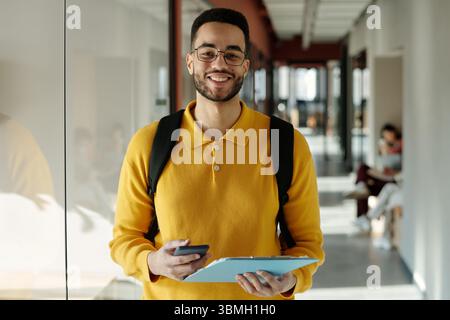 Ritratto di un giovane con la barba che indossa un maglione giallo e occhiali in piedi nello spazio di coworking tenendo in mano lo smartphone e la cartella blu sorridente davanti alla fotocamera Foto Stock