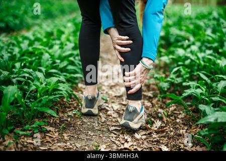Donna che tocca il polpaccio sul sentiero, soffre di dolori alle gambe o crampi durante la corsa nella foresta. Infortunio al fitness Foto Stock