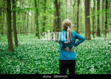Donna che sente dolore alla schiena mentre fa jogging nella foresta. Runner stretching per prevenire il mal di schiena durante l'attività fisica Foto Stock
