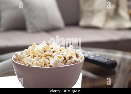 Ciotola con popcorn e telecomando tv su tavolo in vetro vicino al divano Foto Stock