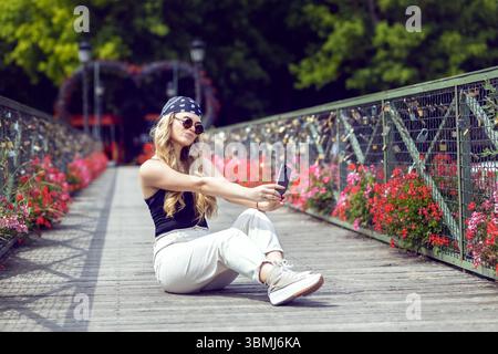 Bella giovane donna bionda hipster che scatta selfie sul romantico ponte dell'amore decorato con fiori e lucchetti turistici Foto Stock
