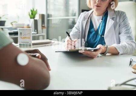 Medico caucasico di mezza età che consulta un paziente adulto di colore con diabete, scrivendo appunti sugli appunti mentre il paziente ha un monitor per il glucosio sul braccio Foto Stock