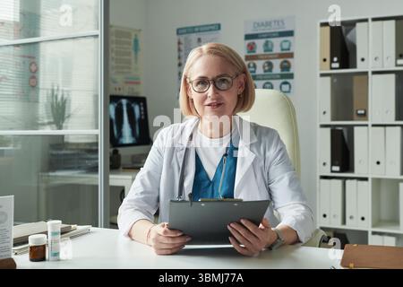 Ritratto della donna caucasica di mezza età dottore seduto alla scrivania tenendo appunti guardando in macchina fotografica in studio medico con manifesti di consapevolezza del diabete Foto Stock