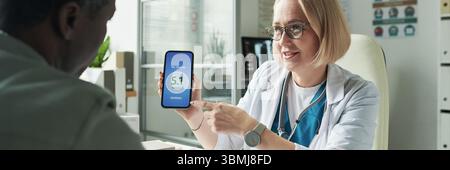 Medico caucasico di mezza età che mostra la lettura digitale del glucosio su smartphone fino a paziente di mezza età di uomo nero durante la consultazione per il diabete in medicina Foto Stock