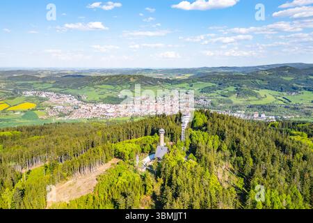 La vista aerea cattura la torre di osservazione di Svatobor circondata da lussureggianti foreste verdi, affacciata sulla città di Susice, annidata nella valle sottostante in una giornata limpida. Foto Stock