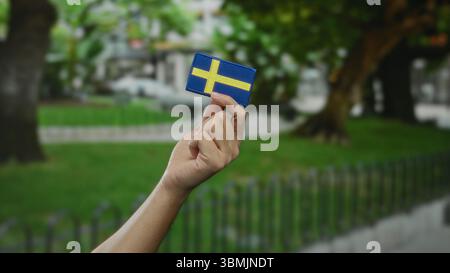 Mano che tiene la bandiera svedese all'aperto in un parco, con l'uomo caucasico visibile, che rappresenta la cultura e l'identità svedesi. Foto Stock