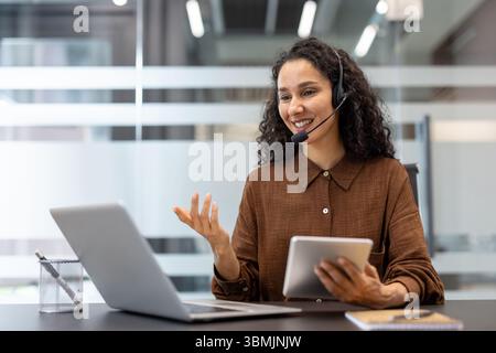 Una donna sorridente che indossa un auricolare partecipa a una videochiamata in ufficio, tenendo in mano un tablet. Foto Stock