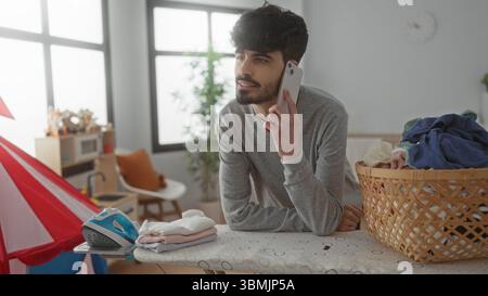Un giovane uomo che parla al telefono mentre stirava in una luminosa lavanderia con un cestino di vestiti e un asse da stiro, mostrando un'atmosfera attraente e informale Foto Stock