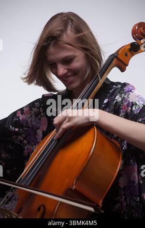 Giovane donna caucasica, 20 anni, sorridente mentre suona il violoncello in studio. Con top floreale nero. Foto Stock