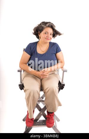 Una donna siede su una sedia con le mani in grembo. Indossa una camicia blu e pantaloni abbronzati. La sedia è piccola, di legno Foto Stock