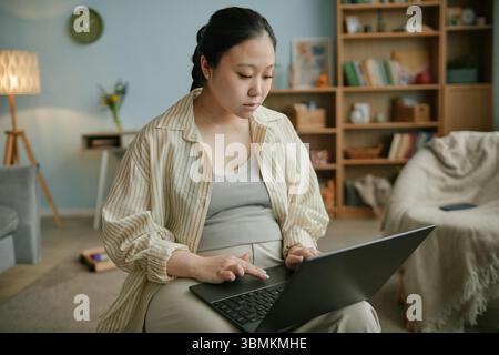 Donna asiatica incinta seduta al chiuso con un laptop, concentrandosi sullo schermo mentre trascorreva il tempo libero a casa, mani sulla tastiera, postura casuale, libreria in background Foto Stock