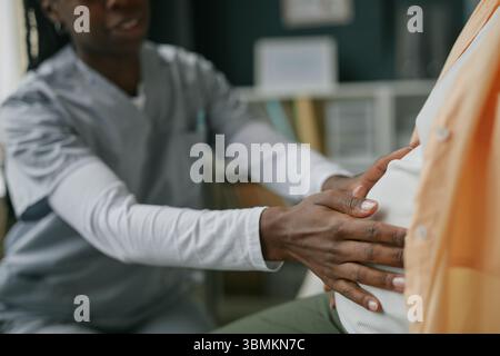 Donna nera che esamina la donna asiatica incinta durante il controllo di cura prenatale, mani che toccano dolcemente l'addome, consultazione medica in ambiente clinico Foto Stock