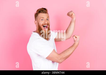 Un uomo allegro con la barba rossa mette in posa una posa flessibile su uno sfondo rosa, mostrando fiducia ed entusiasmo Foto Stock
